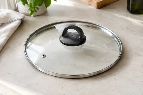 Couvercle rond en verre transparent, rebord en inox et poignée noire posé sur un plan de travail clair